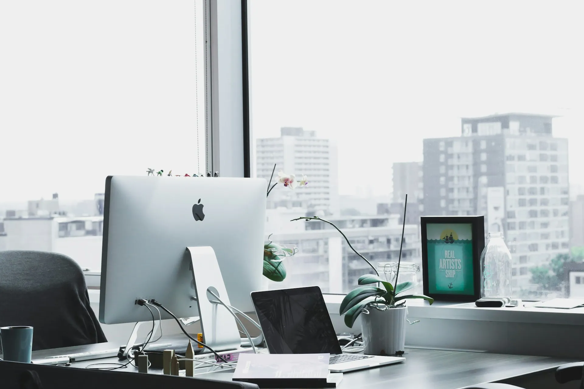 Office desk with a desktop computer and laptop by a city window